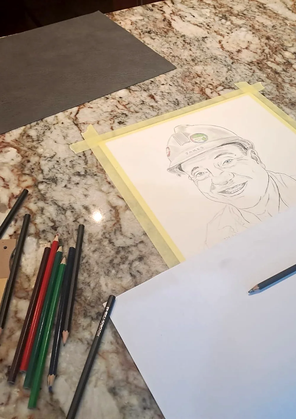 A half-finished pencil portrait of a man in a hard hat, taped to a table beside coloured pencils